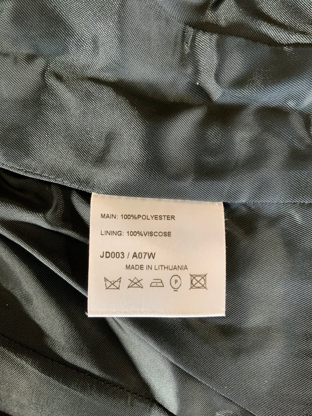 Close-up of a garment's interior lining showing a white care label with text including 'MAIN: 100%POLYESTER', 'LINING: 100%VISCOSE', and 'JD003 / A07W MADE IN LITHUANIA' on dark gray fabric.