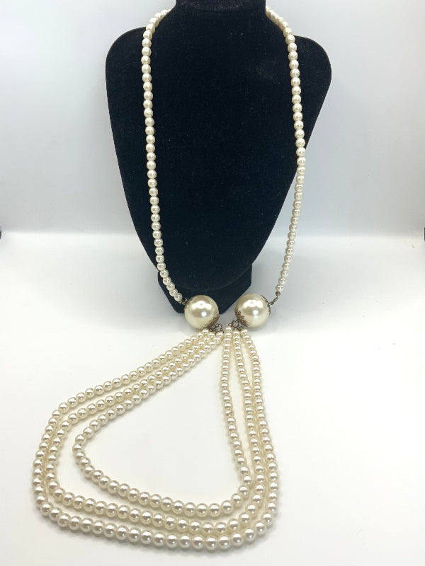 A long pearl necklace displayed on a black mannequin bust against a white studio background. The necklace features multiple strands of small white pearls with two larger round pearls at the center, connected by a thin chain.