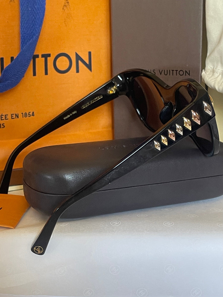 Louis Vuitton sunglasses with black frames, gold-tone rivet details on the temples, and dark lenses, displayed on top of a black case with Louis Vuitton packaging in the background.