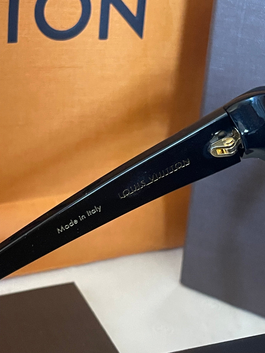 Close-up of a black acetate sunglasses arm with white printed text reading 'LOUIS VUITTON' and 'Made in Italy', resting on a brown cardboard surface.