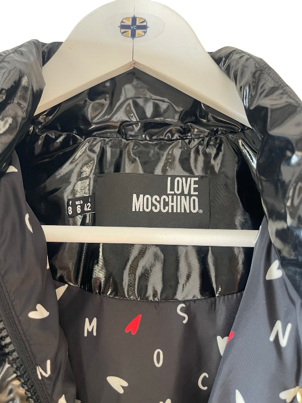 Close-up of a black puffer jacket with a glossy finish, featuring a white 'LOVE MOSCHINO' brand label on the inner collar and a pattern of white and red letters and hearts on the outer fabric. The jacket is displayed on a white hanger against a white background.