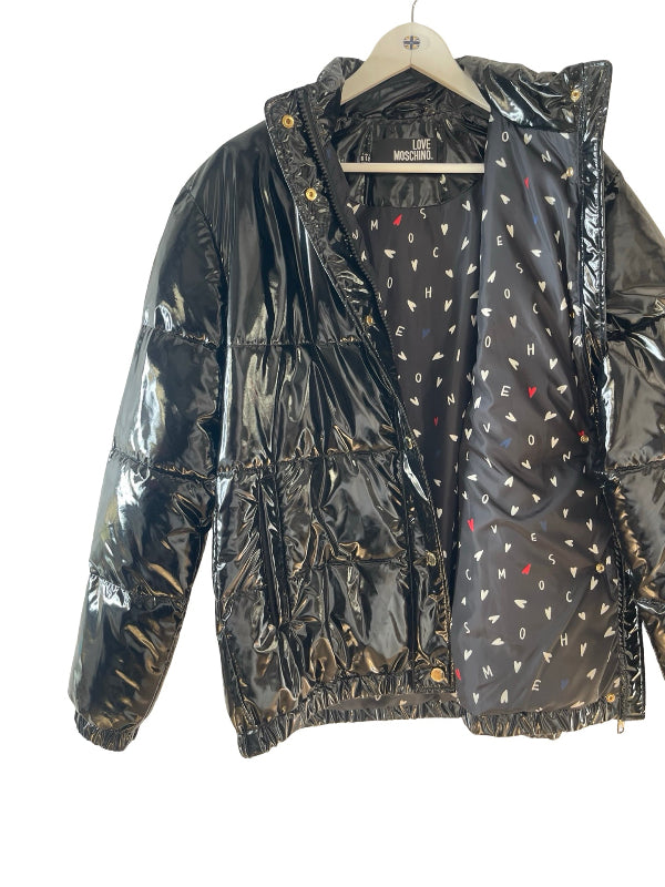 A shiny black puffer jacket with a glossy finish, featuring a patterned black lining with small white, red, heart letter motifs, hanging on a wooden hanger against a white background.