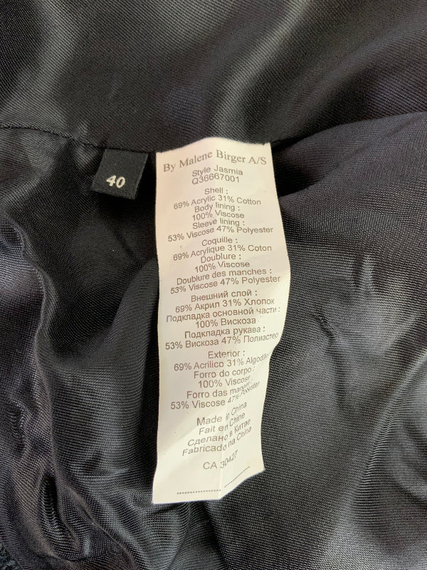 Close-up of the inside collar and care label of a black garment. The white paper label reads 'By: Malene Burger A/S' and shows material composition details.