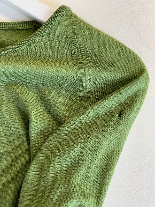 Close-up of an olive green knit top on a white hanger, showing the ribbed collar, shoulder seam, and sleeve texture against a plain off-white background.