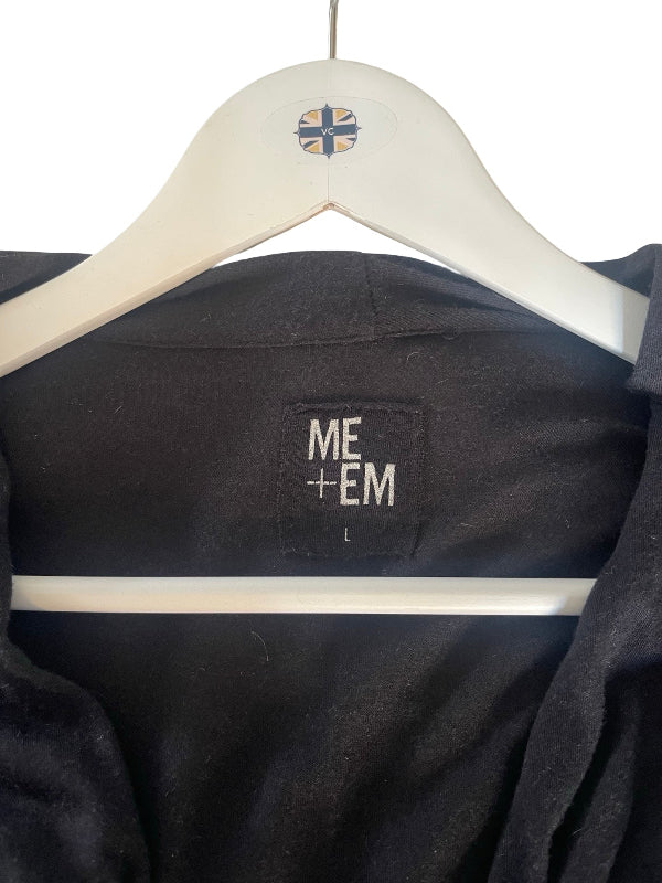 A close-up of a black garment hanging on a white hanger against a white background. The inner neck label reads 'ME+EM', and a small ME+EM logo is visible on the hanger.