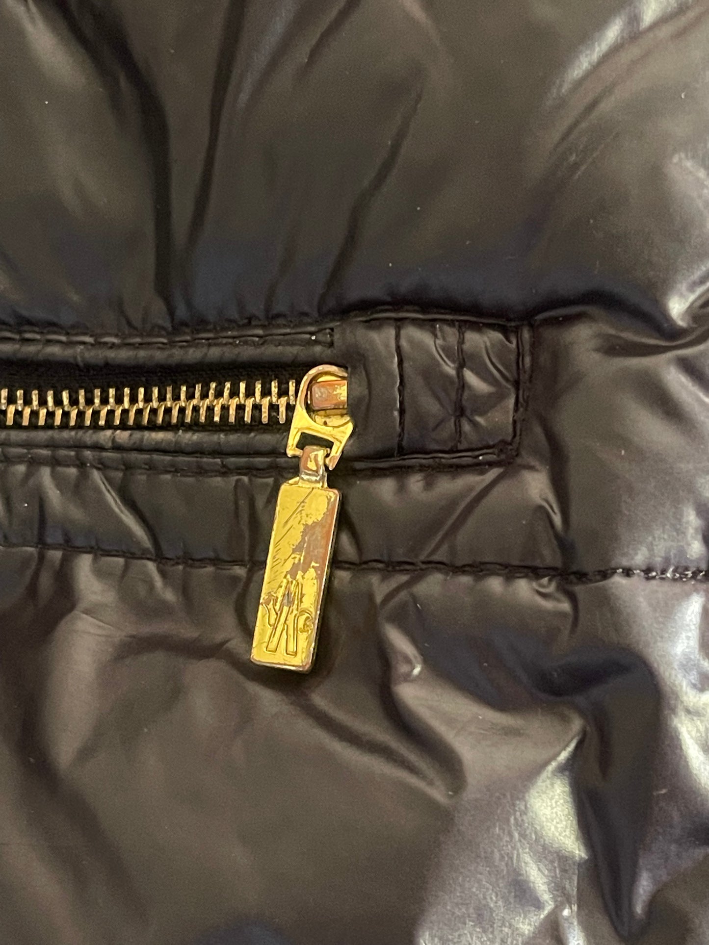Close-up of a black quilted puffer jacket showing a gold-tone metal zipper pull with an engraved logo.