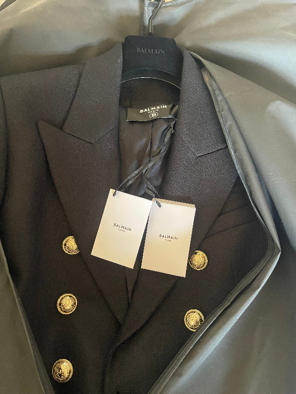 A Balmain double-breasted blazer in dark black, featuring gold buttons and white Balmain tags, inside a black garment bag.