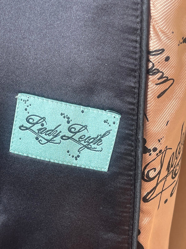 Close-up of a black satin garment with a mint green fabric patch reading 'Lady Leigh' in black script, next to a peach-colored patterned fabric panel.
