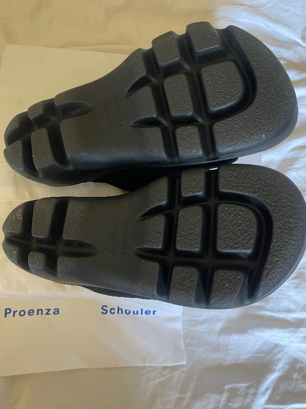 A pair of black slide sandals shown from the sole side, resting on a white fabric surface with a white paper insert printed with the names 'Proenza' and 'Schouler'.