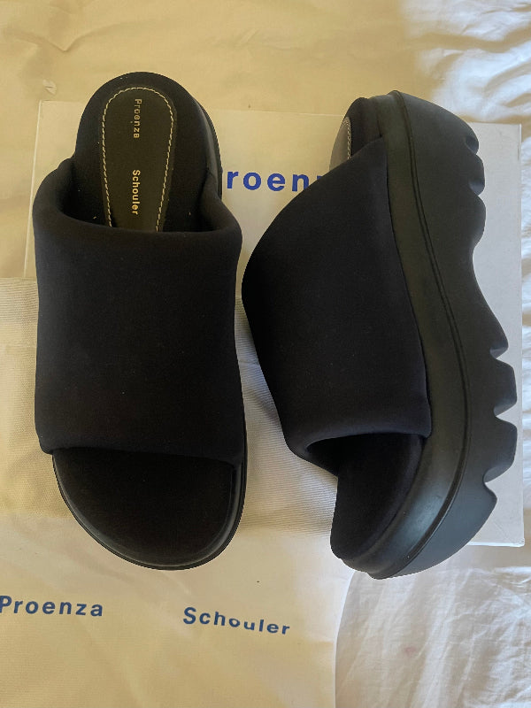 A pair of black platform slide sandals with wide straps, shown on top of white Proenza Schouler packaging. One sandal is viewed from above, and the other shows the side profile of its chunky sole.