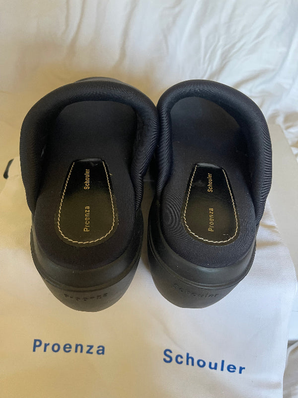 A pair of black Proenza Schouler clogs shown from the sole side, placed on a white fabric surface next to their branded white dust bag.