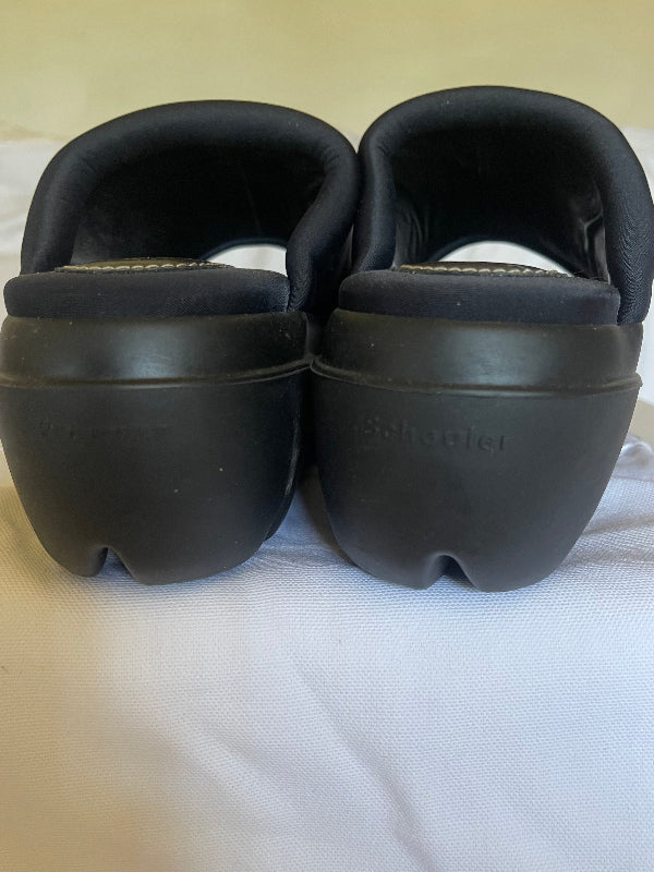 A pair of black platform clogs with thick soles, photographed from the back on a white fabric surface.