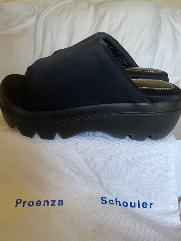 A single black platform slide sandal with a chunky lug sole, displayed on a white fabric surface with the text 'Proenza Schouler' printed below it.