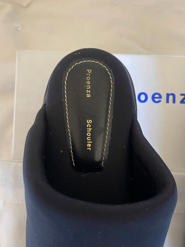 Close-up of the heel area of a black slip-on shoe, showing the insole with 'Proenza Schouler' printed in gold and white contrast stitching. A white shoebox with blue 'Proenza' lettering is partially visible in the background.