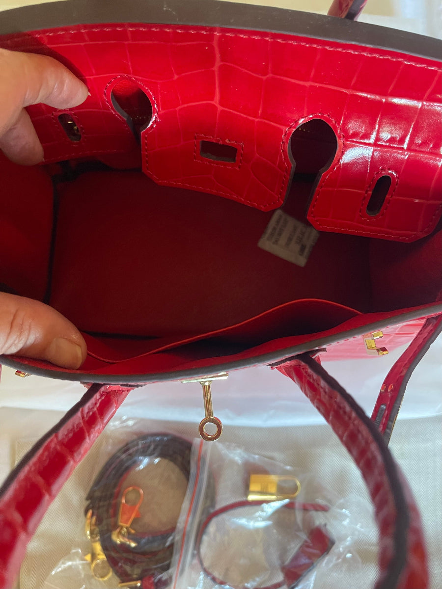 Close-up of the interior of a red crocodile-embossed leather handbag, showing the lining, interior pockets, and a small attached product tag. A hand is holding the bag open, and the bag's strap and lock hardware are visible below.