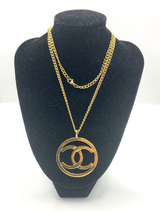 A gold-tone double chain necklace with a large interlocking CC pendant, displayed on a black velvet jewelry bust against a white background.