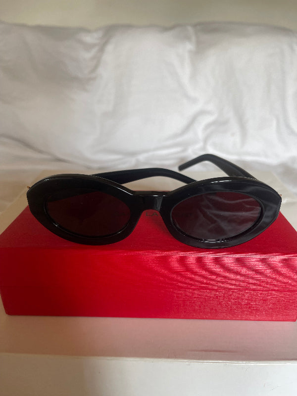 Black round-frame sunglasses with dark lenses resting on a red box next to a white fabric surface.