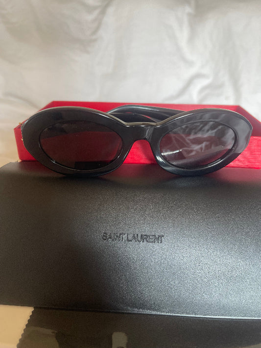 Black oval-frame sunglasses with dark lenses resting on a red box next to a black Saint Laurent case, with a white fabric background.