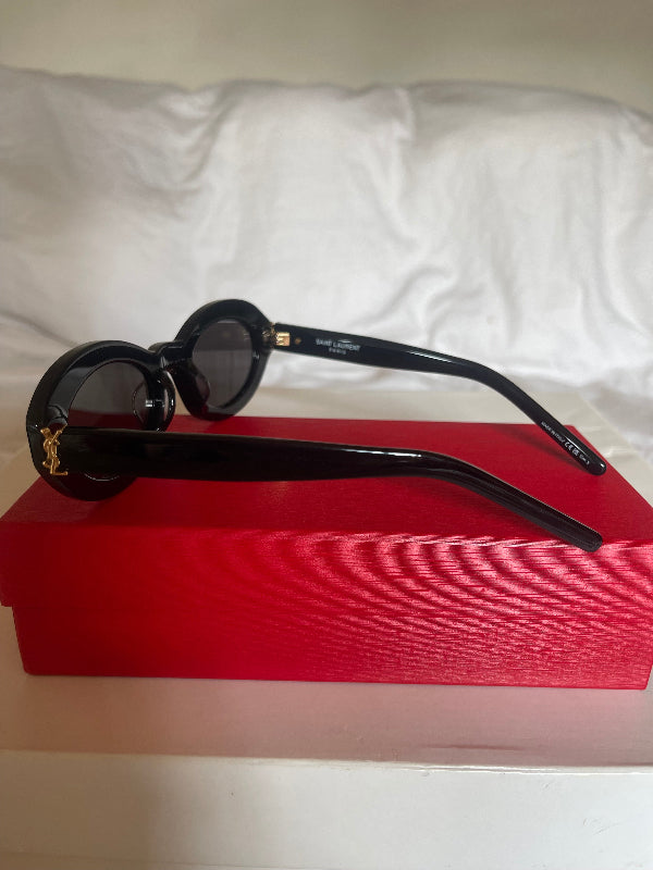 Black round-frame sunglasses with dark lenses and gold-tone logo details, resting on a red rectangular box against a white fabric background.