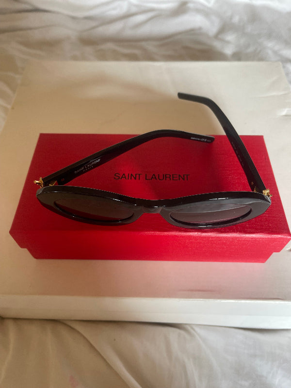 Saint Laurent round sunglasses with black frames and dark lenses, resting on a red branded case against a white box, with white fabric visible around it.