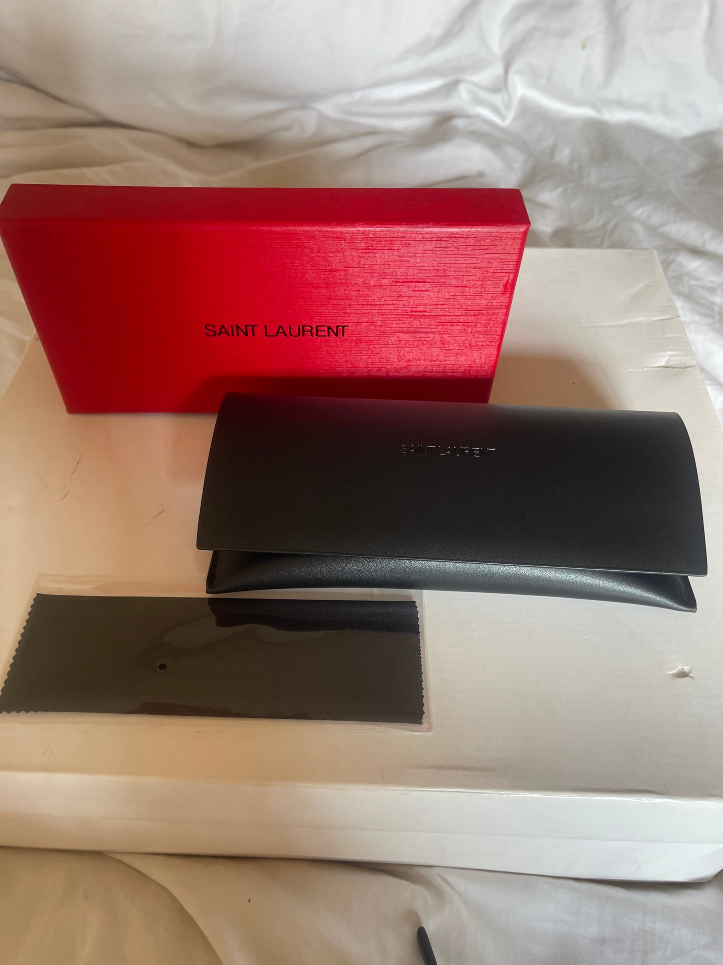 Saint Laurent eyewear accessories including a red Saint Laurent gift box, a black leather case, and a black cleaning cloth, displayed on a white surface.