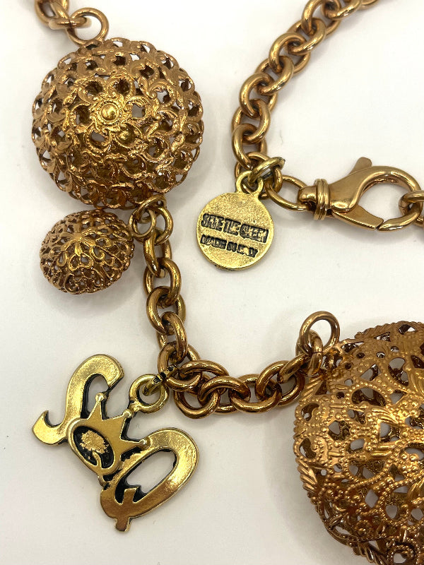 Close-up of a gold-tone charm bracelet featuring textured filigree ball charms, a script letter charm, and a round engraved tag on a white background.