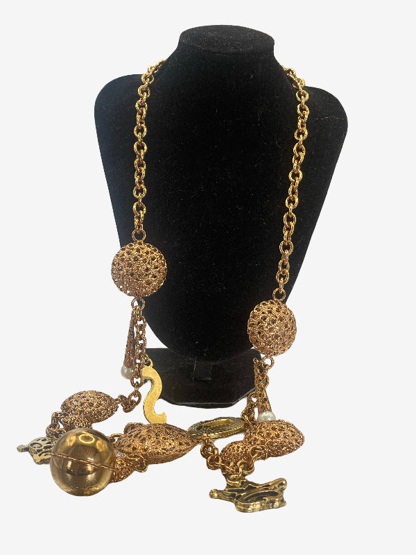 A gold-toned statement necklace displayed on a black velvet jewelry bust. The necklace features a chunky chain, two large textured spherical beads, a small gold bird charm, and several dangling organic-shaped charms, including a round gold bead and a small crown figure.