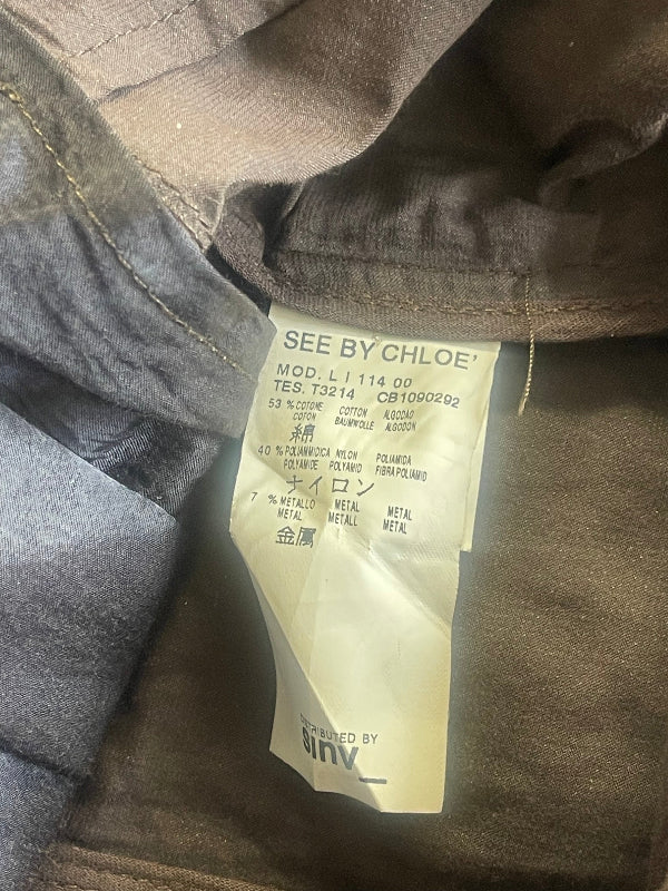Close-up of the inside neck area of a See By Chloe garment, showing a cream paper size and care label attached to dark brown and gray textured fabric.