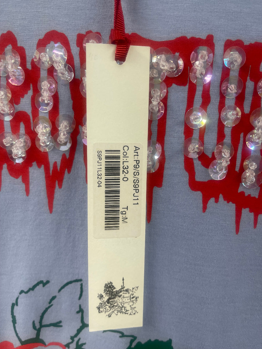 A close-up of a garment or textile featuring a red abstract pattern on a light blue-gray base, decorated with rows of clear, round sequins. A white paper hangtag is laid horizontally across the center, displaying product code text and a barcode.