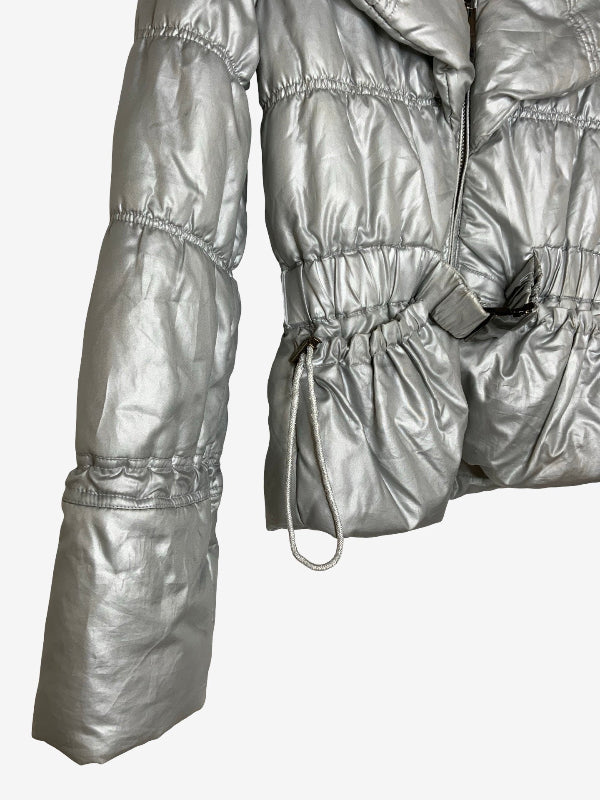 A close-up of a metallic silver puffer jacket with horizontal quilted baffles, elasticized cuffs, and a drawstring waist detail, shown against a plain light gray background.