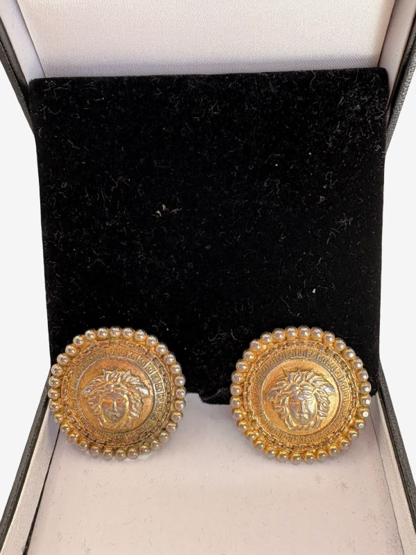 A pair of round gold-tone earrings displayed in an open jewelry box. Each earring features an embossed Medusa-style face surrounded by a border of small pearls, set against a black velvet interior.