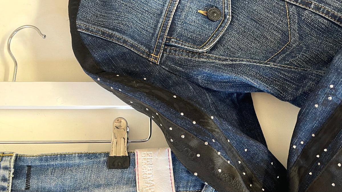 A close-up of a blue denim jacket with black faux-leather trim and silver rhinestone embellishments, hanging on a white hanger with other jeans below it.