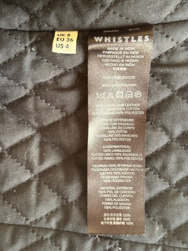 Close-up of a garment's interior label reading WHISTLES, with a small yellow size sticker above it, photographed against a quilted black fabric background.