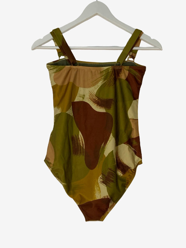 A one-piece swimsuit with a camouflage pattern in olive green, brown, tan, and cream, featuring shoulder straps and a scoop neckline, hanging on a white hanger against a light gray background.