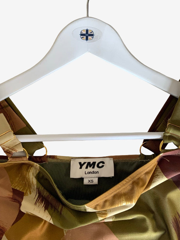 A close-up of a YMC London garment hanging on a white hanger against a plain white background. The garment has a satin-like finish with a pattern of olive green, gold, and brown, and a white size label reading 'YMC London' and 'XS'.