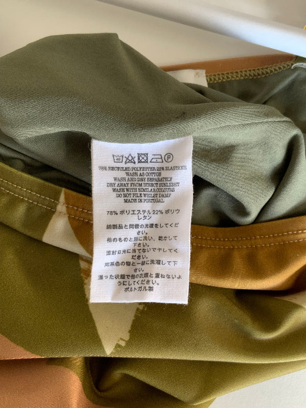 Close-up of a garment's interior label sewn onto olive green fabric with a gold zipper trim, photographed on a light surface.
