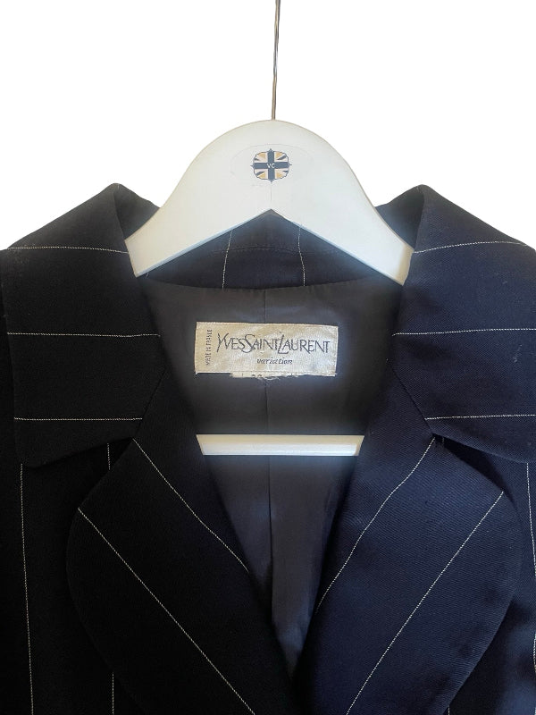 A close-up of a black Yves Saint Laurent 1940 Edition blazer with thin white pinstripes, hanging on a white wooden hanger against a white background.