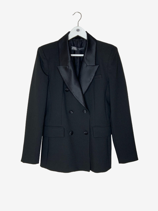 A black double-breasted blazer with satin lapels, hanging on a white hanger against a plain white background.