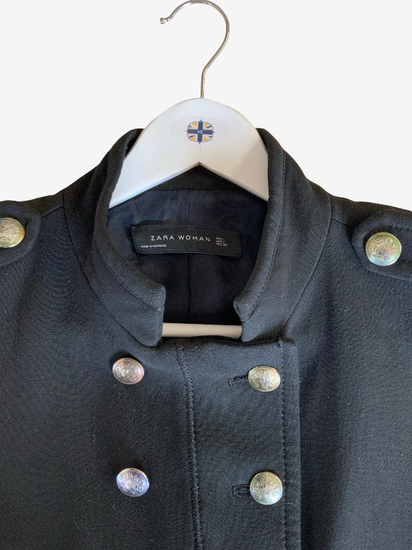 A close-up of a Zara Woman black military-style jacket hanging on a white hanger with a small Union Jack flag logo. The jacket features a high collar, double-breasted silver buttons, and decorative metallic shoulder buttons.