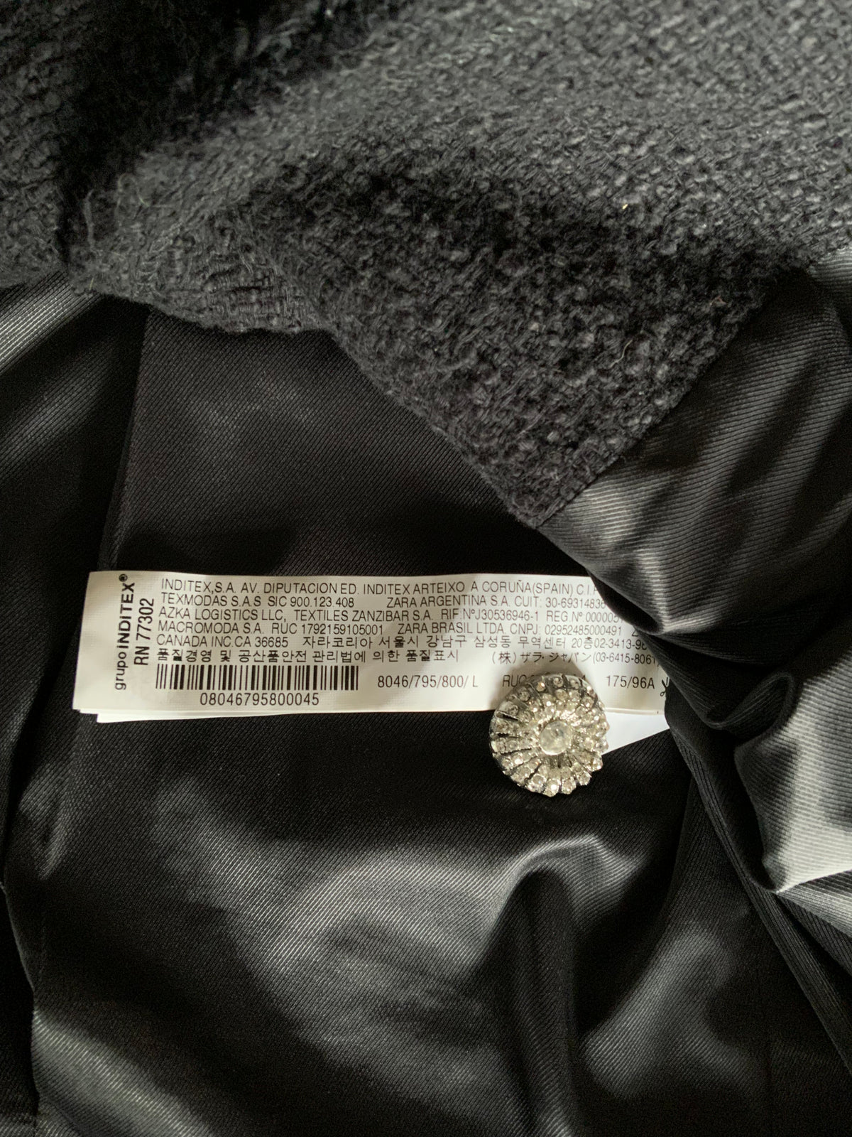 Close-up of a black garment interior showing a white sewn-in size and care label, a small round silver-tone embellished button, and dark textured fabric.