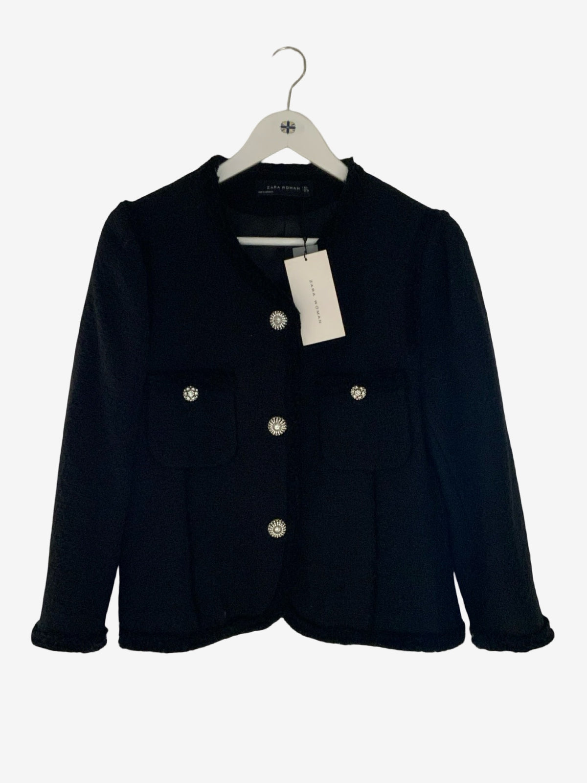 A black cropped jacket with silver-tone buttons and decorative brooch details on the chest pockets, hanging on a white hanger with a paper tag against a light gray background