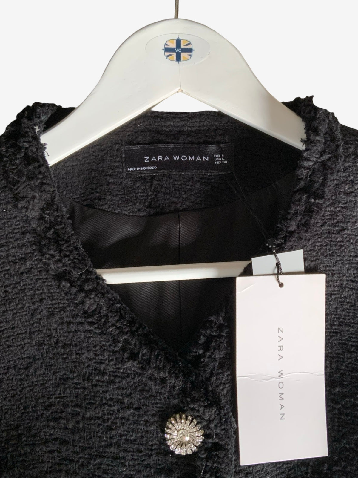 A close-up of a Zara Woman black textured tweed jacket on a white hanger, featuring a black satin lining, a decorative crystal-embellished button, and a white Zara Woman brand hangtag.