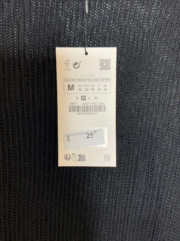 A size tag for a knit garment hanging against dark textured fabric. The tag shows size information  M, a barcode, a price sticker reading £25.99 and the brand name