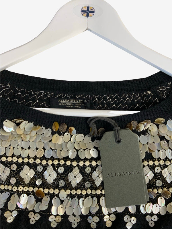 A close-up of a black knit sweater decorated with silver, white, and gold sequins, hanging on a white hanger with an All Saints tag.