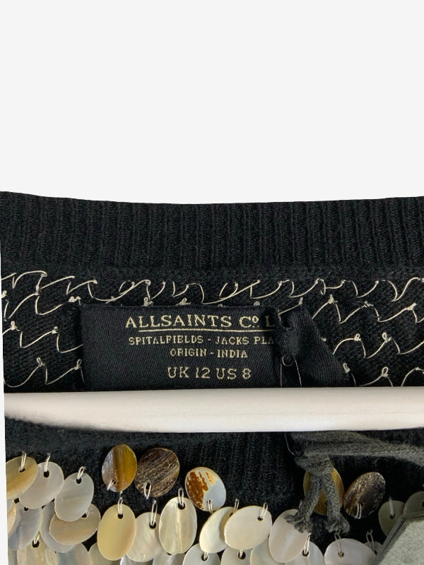 Close-up of a black ribbed knit garment label reading ALLSAINTS CO, with decorative gold embroidery and a row of shell-like sequins along the hem.