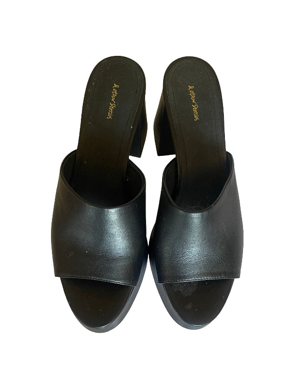 A pair of black leather slide mules with block heels, shown from a top-down view on a white background. The brand name 'and other stories' is printed in gold on the insoles.