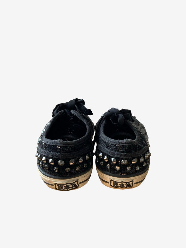 A pair of black low-top sneakers with silver metal studs and distressed detailing, shown from the back on a light gray background.