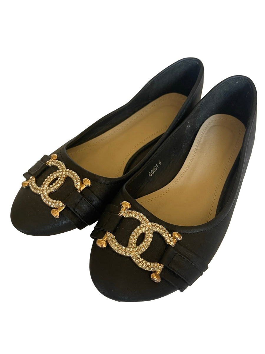 A pair of black leather ballet flats featuring a decorative gold-tone metal buckle embellished with clear rhinestones, displayed on a clean white background.
