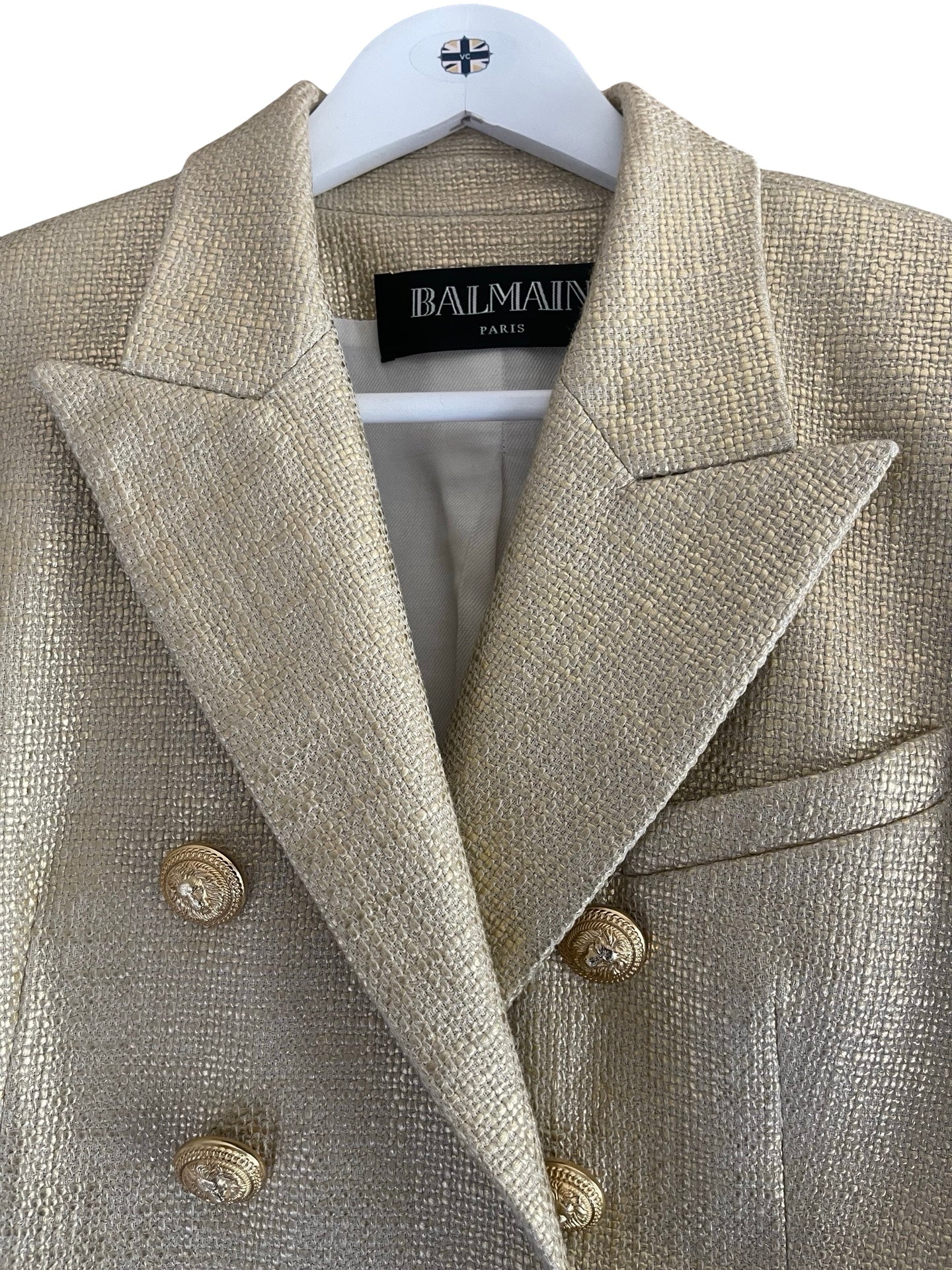 A close-up of a Balmain metallic champagne-colored double-breasted blazer on a white hanger, showing the textured tweed fabric and gold-tone buttons.