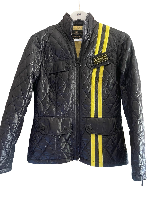 A black quilted Barbour jacket with two vertical yellow stripes on the right side and a yellow Barbour logo patch on the left chest, shown hanging on a white hanger against a white background.